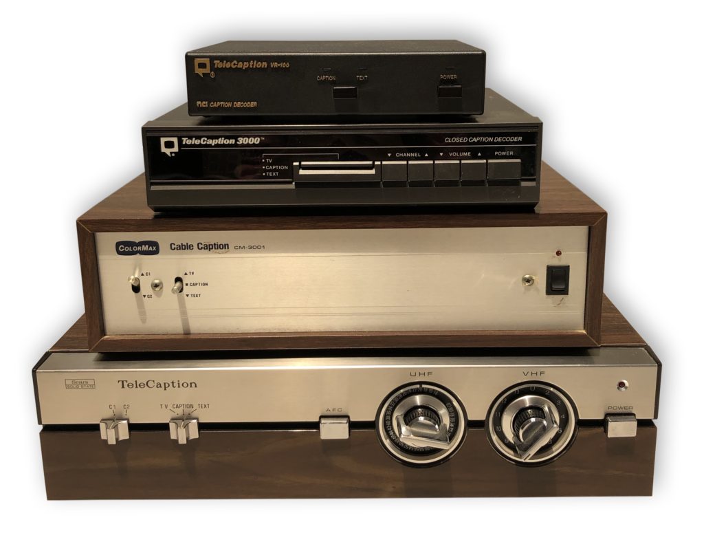 Four closed caption decoder boxes stacked on top of each other. The bottom two boxes have a 1970s wood design. The top two boxes have a more modern and streamlined design.