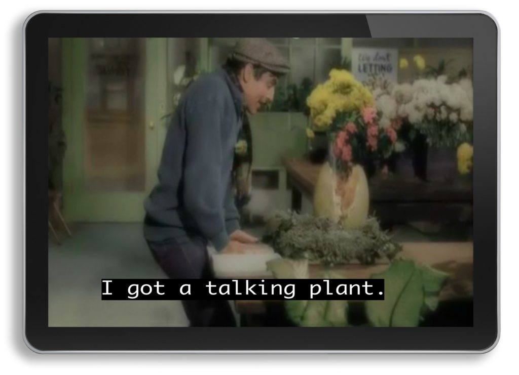 In the tablet, there is a scene from the movie "Little Shop of Horrors." A white man who dresses in a dark blue coat and jeans with a duckbill cap is standing. He looks down at a plant on the table. The caption, in high-definition white on a black background, reads "I got a talking plant."