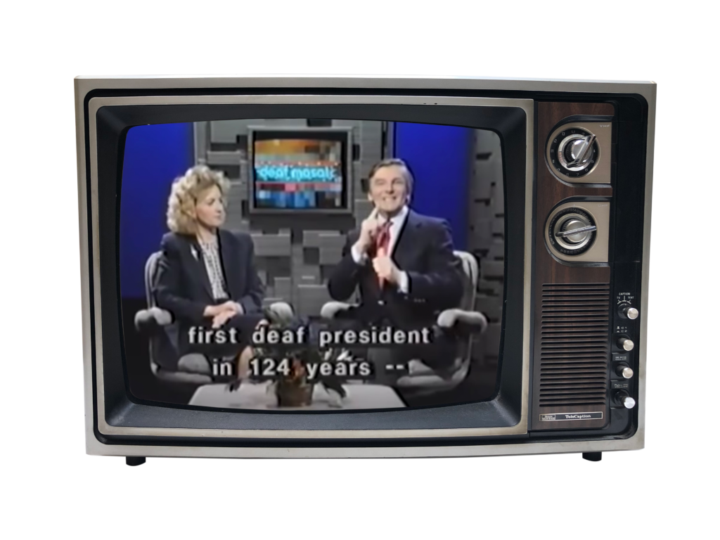 There are a man and a woman in this old TV with two dials for changing program and volume. They are white people with formal suits, in the Deaf Mosaic program. They are sitting on the chair and facing the camera. The woman is watching the man sign. This man who has a smile on his face is telling the story of Deaf President Now. The caption reads "first deaf president in 124 years --"   